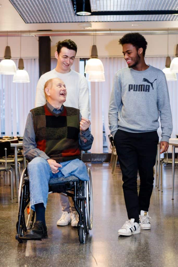 Kaksi nuorta kuljettaa pyörätuolissa olevaa senioria